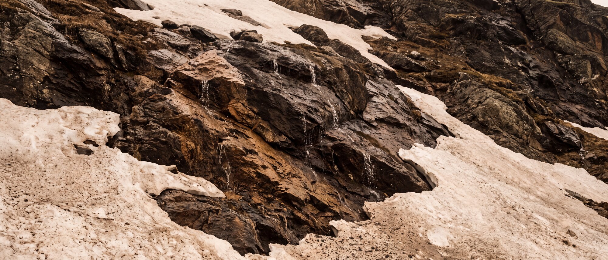 Versante bagnato di roccia scura e neve vecchia, ripreso da vicino senza cielo.